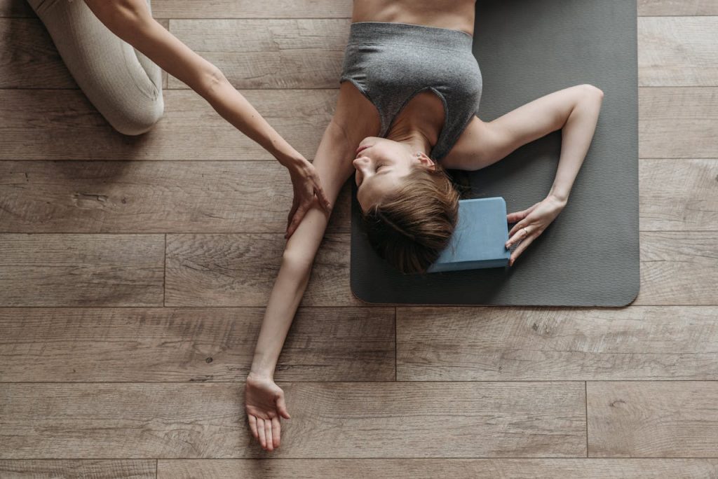 Woman lying on yoga mat being assisted in stretching routine by a trainer indoors.